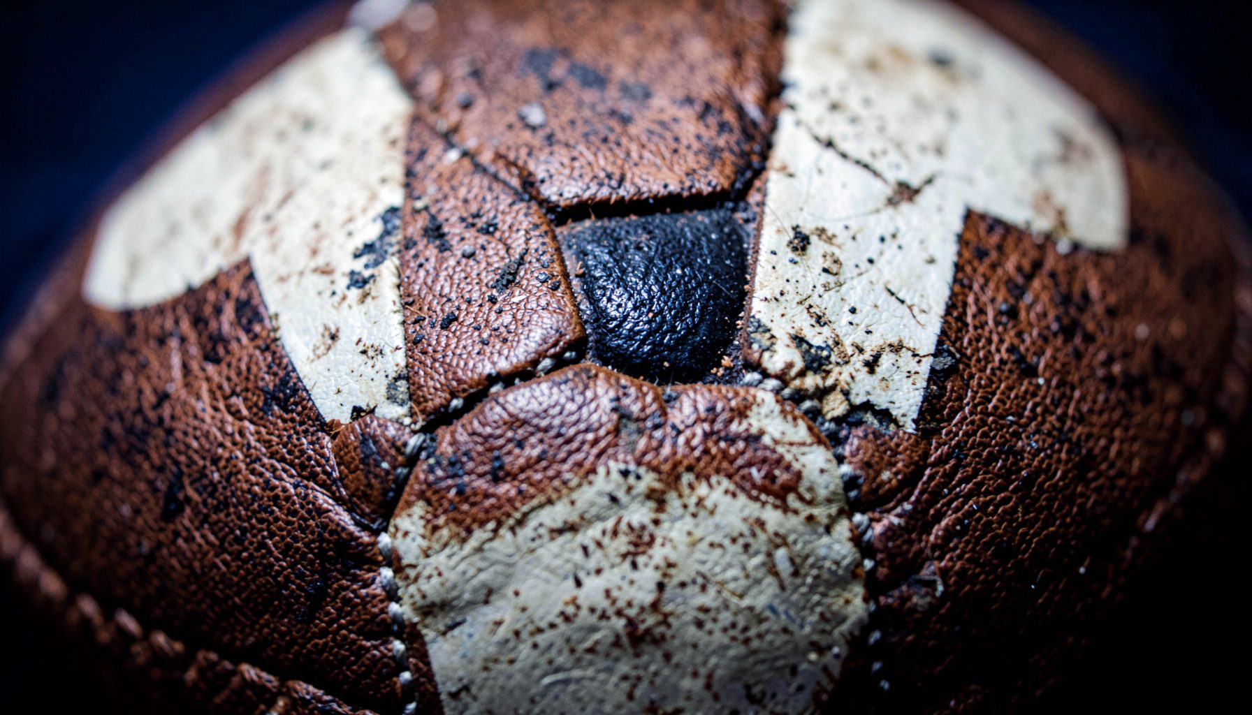 Extreme close-up of a worn leather football surface, scuffed white panels and black pentagon stitching, rich brown tones