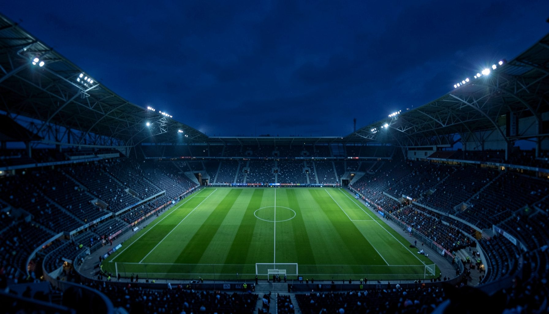 Wide aerial view of a floodlit football stadium at dusk, deep navy sky with scattered floodlight glow, green pitch centered below