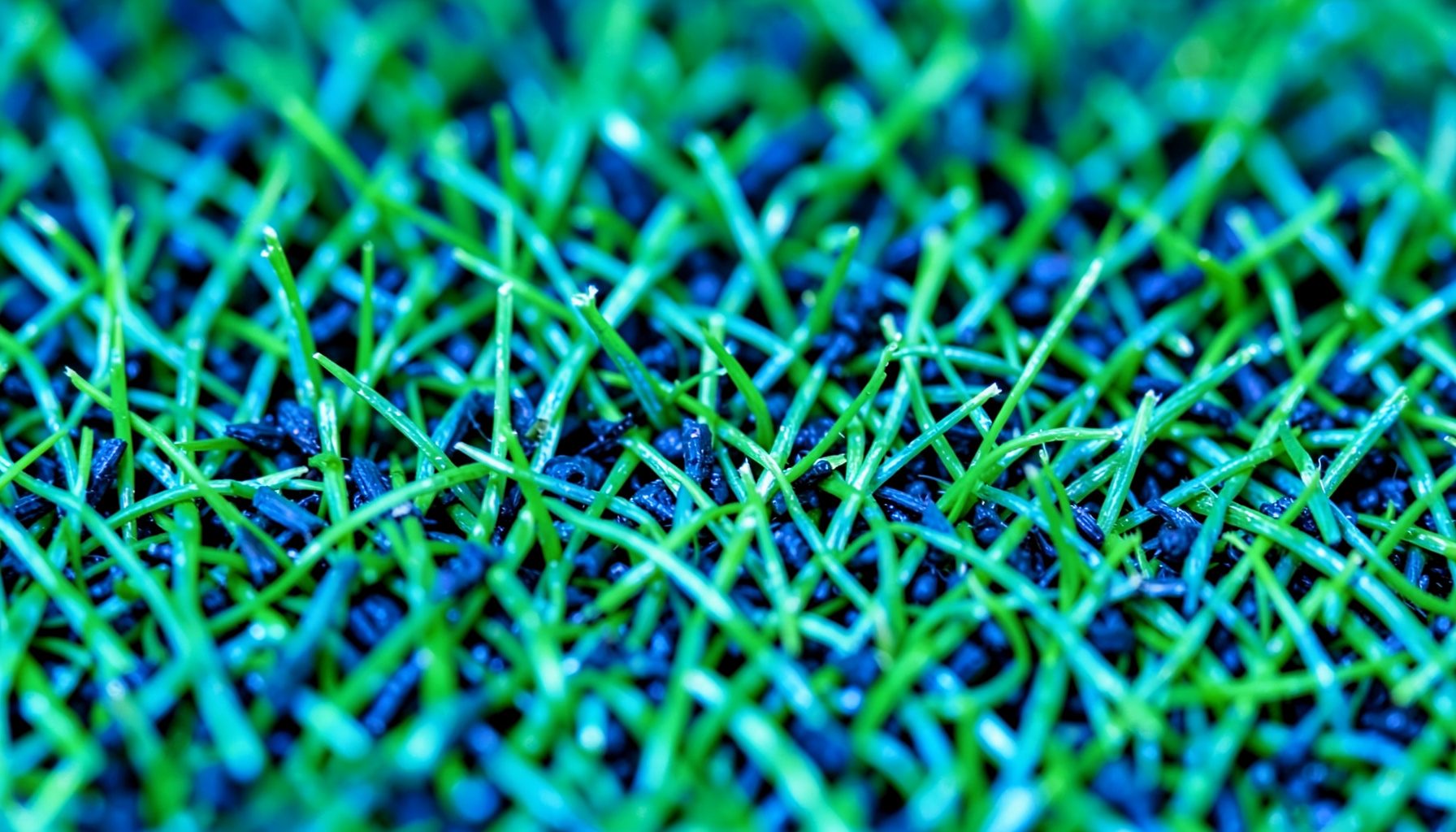 Extreme close-up of green artificial turf fibers with black rubber infill granules scattered between blades, bright and textured