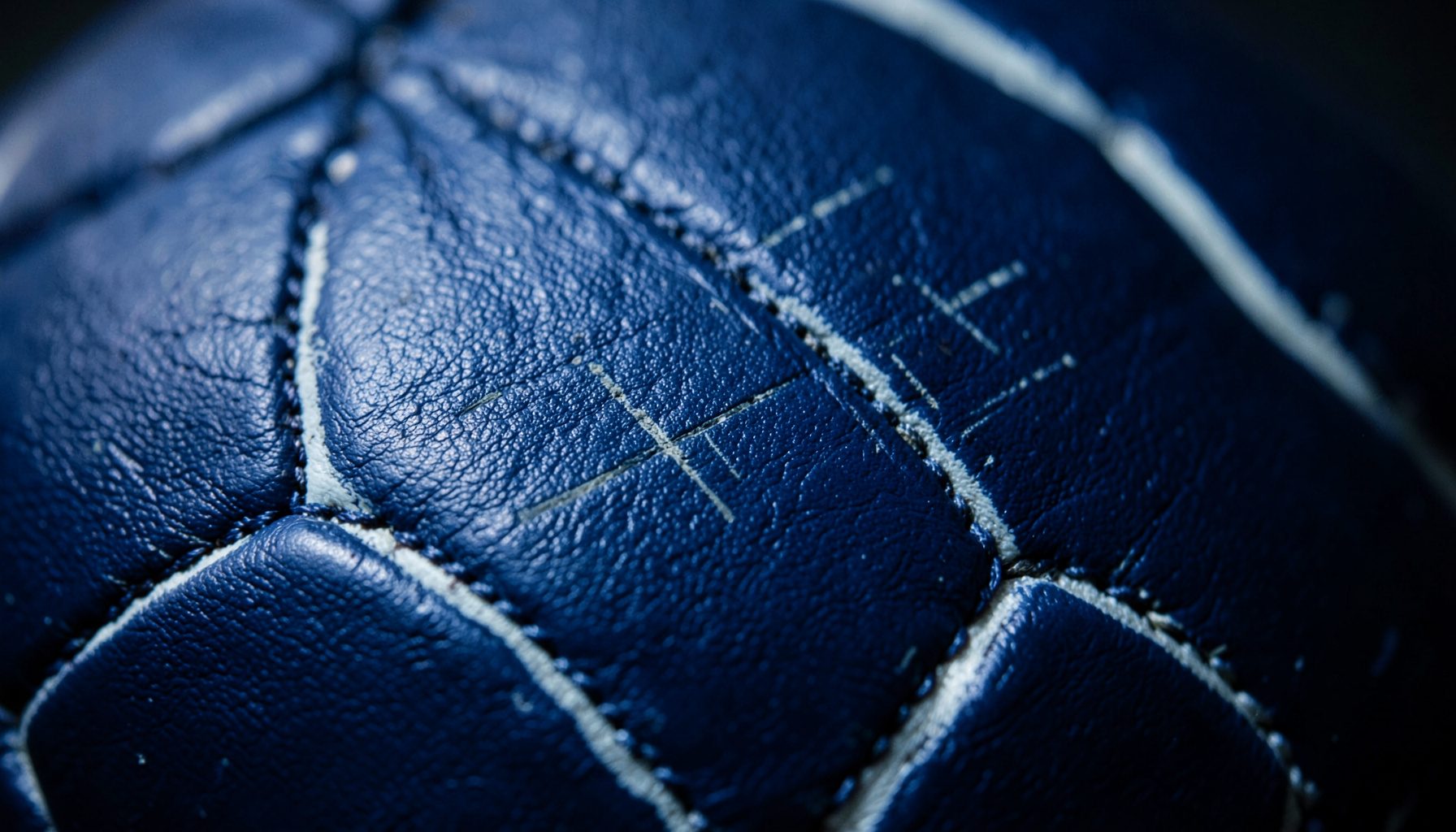 Extreme close-up of a football's stitched leather surface, deep navy and white hexagonal panels with subtle scuff marks